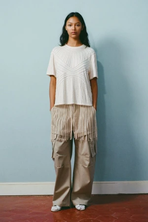 Model standing against a blue wall wearing a cream textured knit t-shirt and beige cargo pants with fringe detailing.