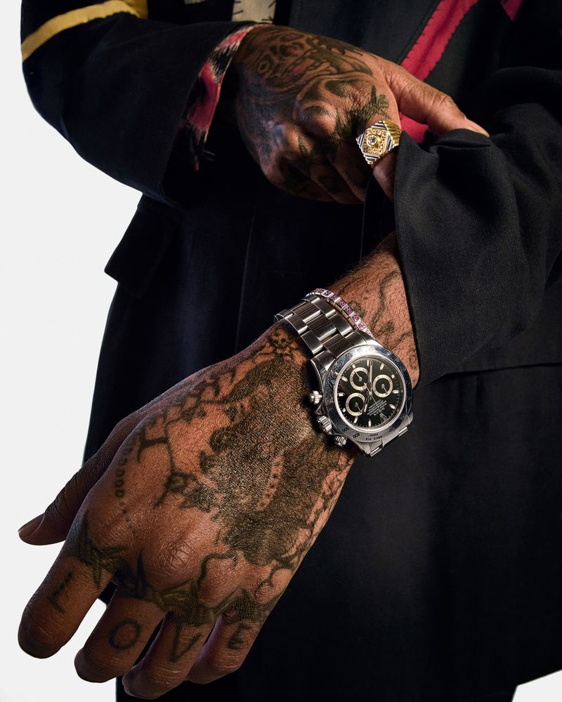 Close-up of a man's arm and hand with a wrist watch, gold ring, and tattoos, set against a clean, white backdrop.