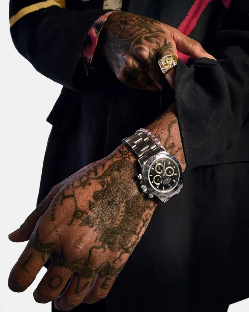 Close-up of a man's arm and hand with a wrist watch, gold ring, and tattoos, set against a clean, white backdrop.