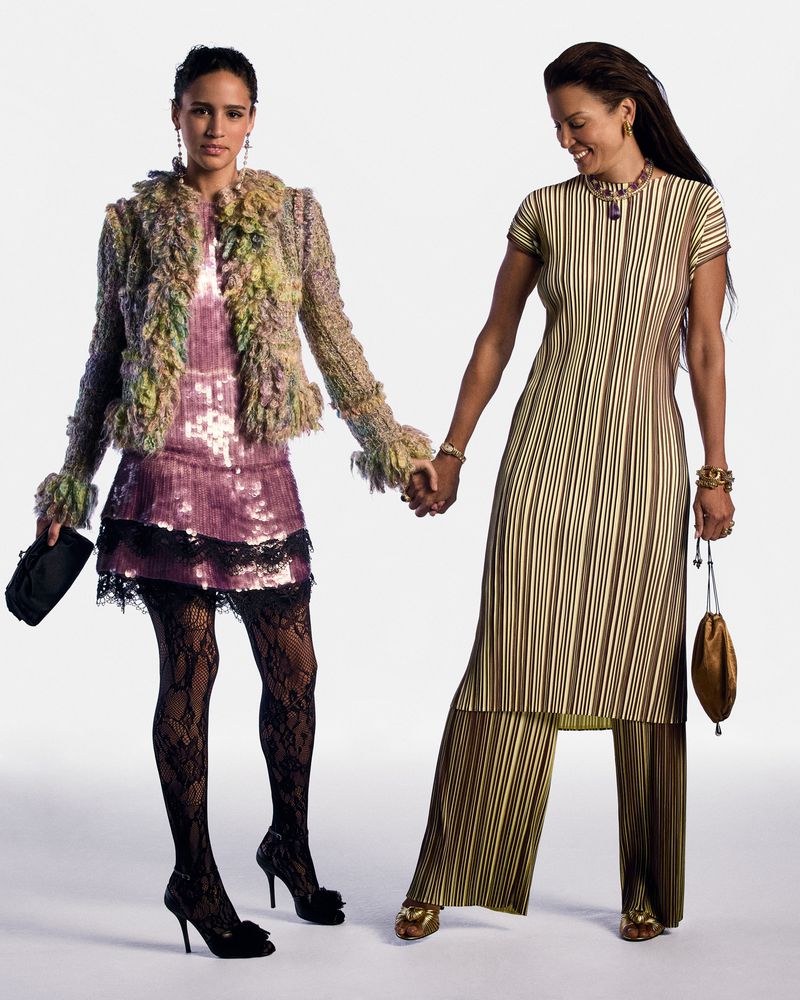 Two adult women holding hands in studio. Woman on the left wears sequin dress with furry jacket and heels, right wears striped tunic and pants.