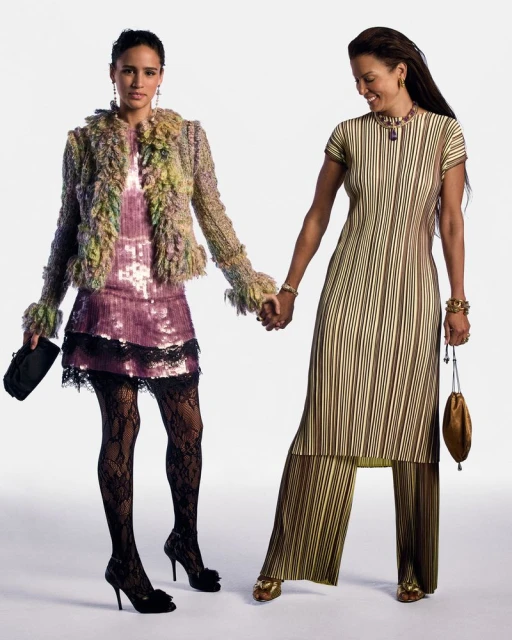 Two adult women holding hands in studio. Woman on the left wears sequin dress with furry jacket and heels, right wears striped tunic and pants.
