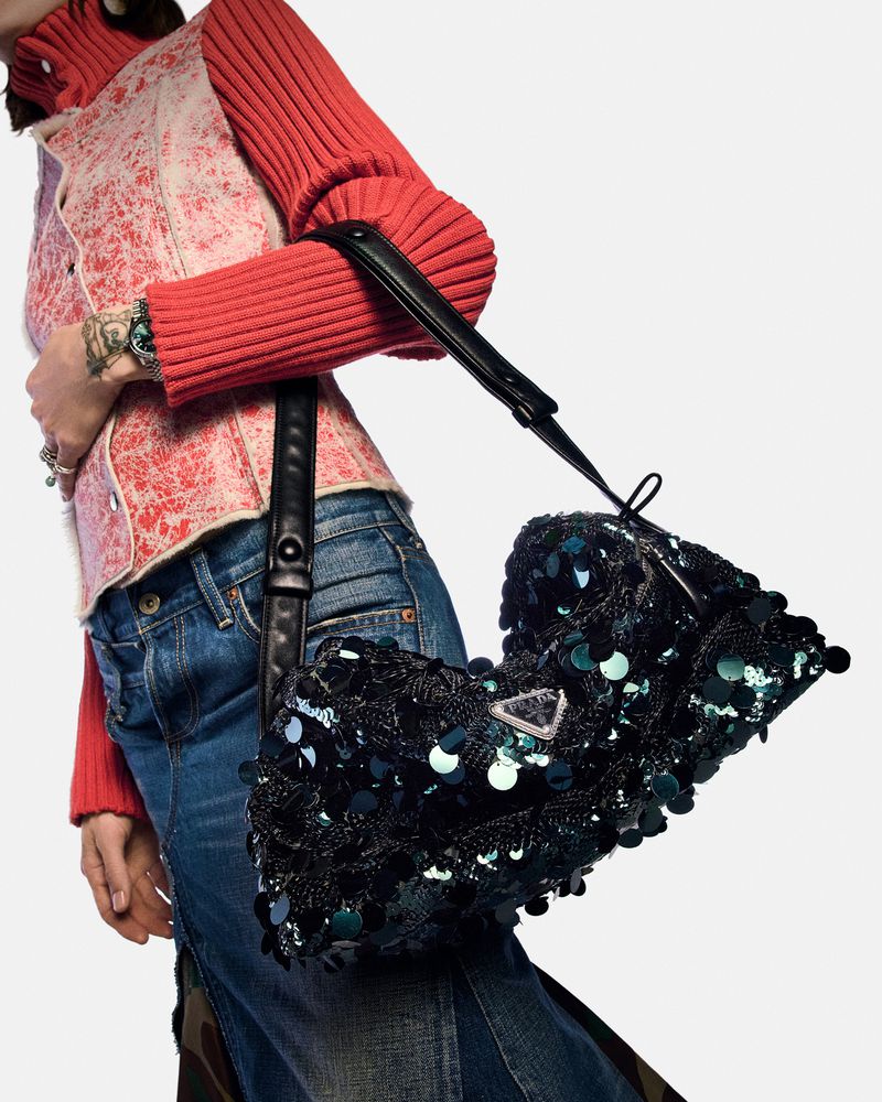 A woman is shown from the waist down, carrying a black, sequined Prada bag with a shoulder strap, against a white background.