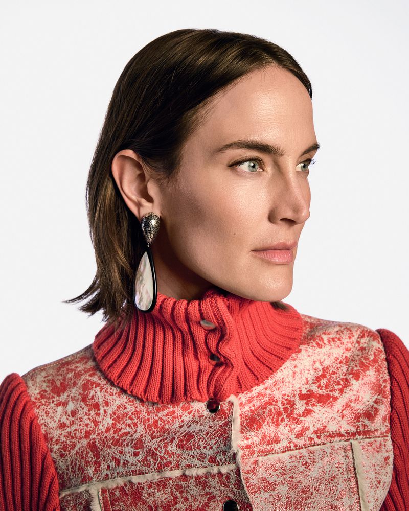 Close-up portrait of a woman with short brown hair wearing an embellished red jacket and large teardrop earrings against a white backdrop.