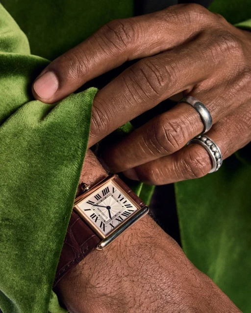 Close-up of a dark-skinned hand adorned with silver rings and a gold and brown watch, resting against a green velvet surface.