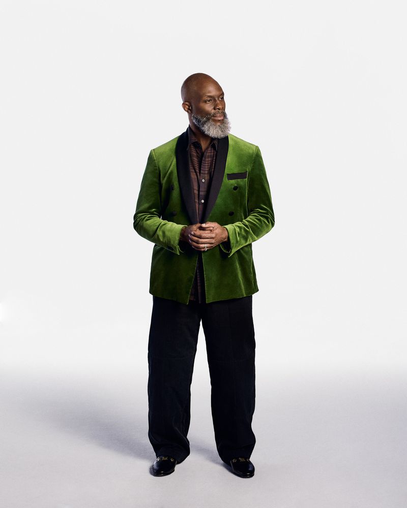 Full body studio shot of a mature Black man with a beard in a green velvet blazer, black shirt, and black pants looking away.