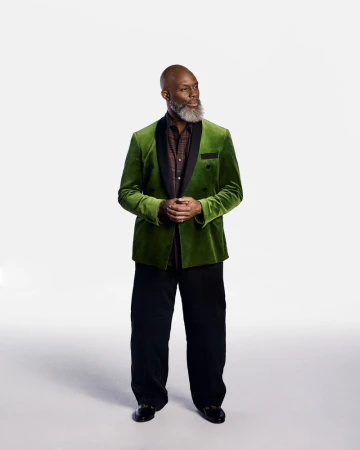 Full body studio shot of a mature Black man with a beard in a green velvet blazer, black shirt, and black pants looking away.