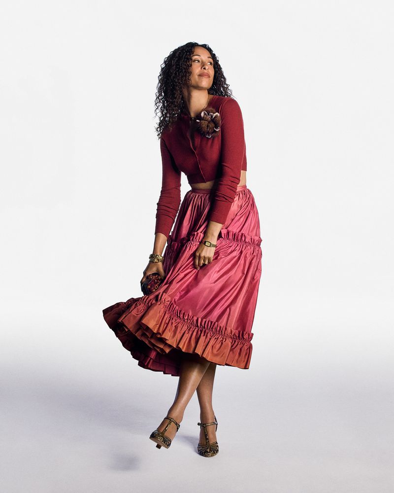 Adult woman in a red cropped cardigan and layered pink skirt stands in a studio with curly hair and jewelry looking upwards.