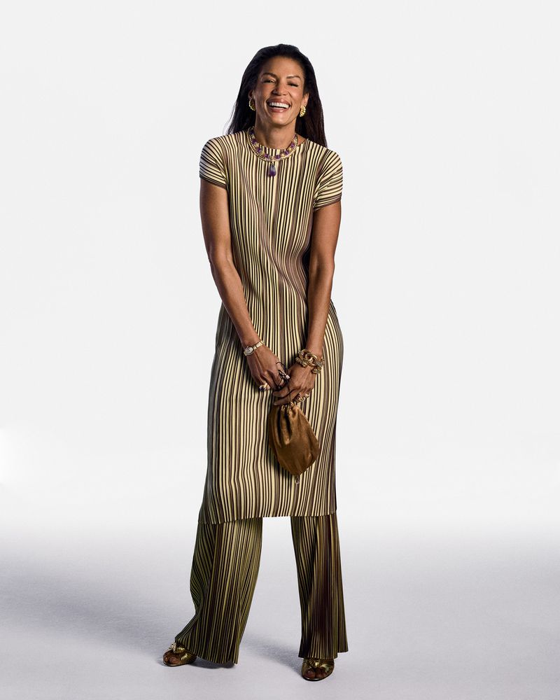 Smiling adult female stands in a photo studio wearing a striped olive-green matching top and pants with gold jewellery and gold heels.