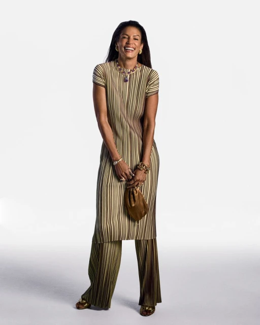 Smiling adult female stands in a photo studio wearing a striped olive-green matching top and pants with gold jewellery and gold heels.