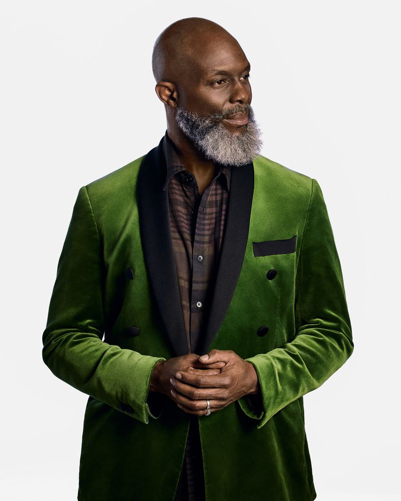 Portrait of a mature Black man in a green velvet suit with a patterned shirt, standing against a bright white backdrop, looking off-camera.