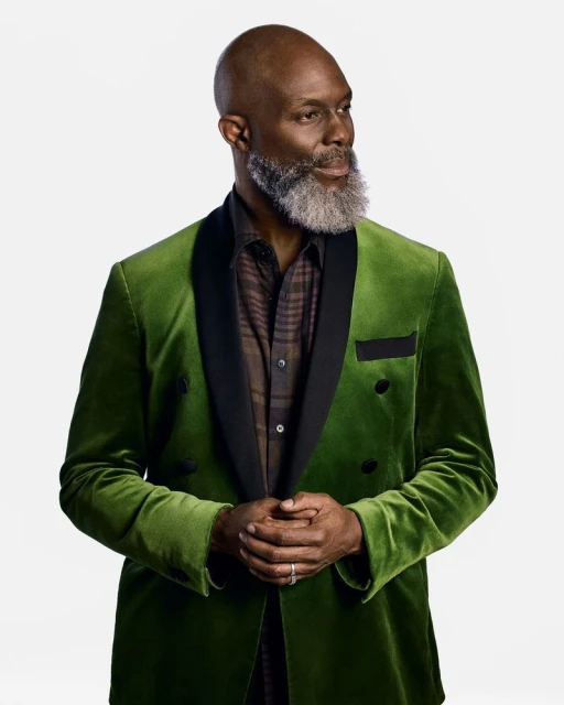 Portrait of a mature Black man in a green velvet suit with a patterned shirt, standing against a bright white backdrop, looking off-camera.