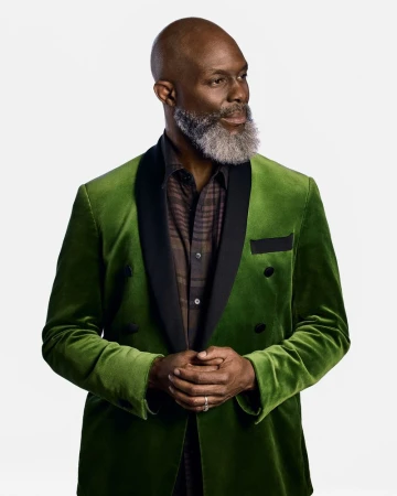 Portrait of a mature Black man in a green velvet suit with a patterned shirt, standing against a bright white backdrop, looking off-camera.