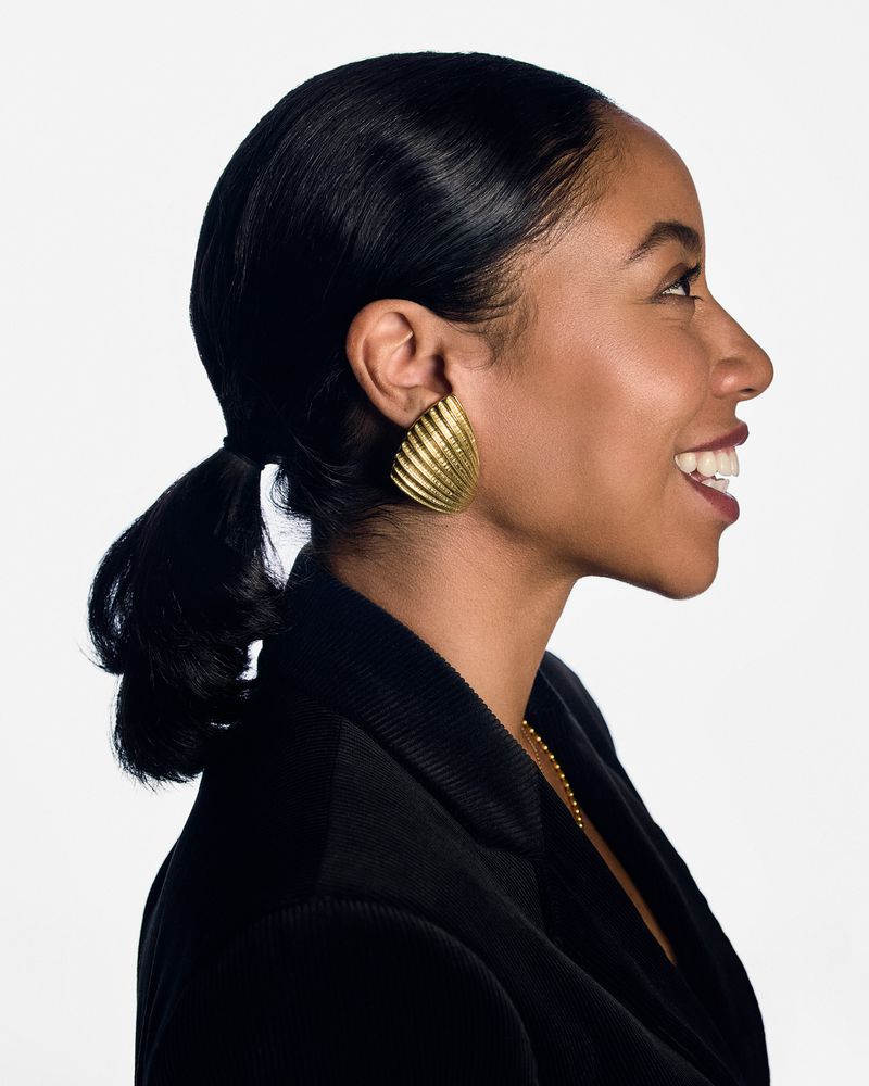 Profile shot of a smiling adult woman with dark hair in a ponytail wearing a black jacket and gold earrings against a white backdrop.