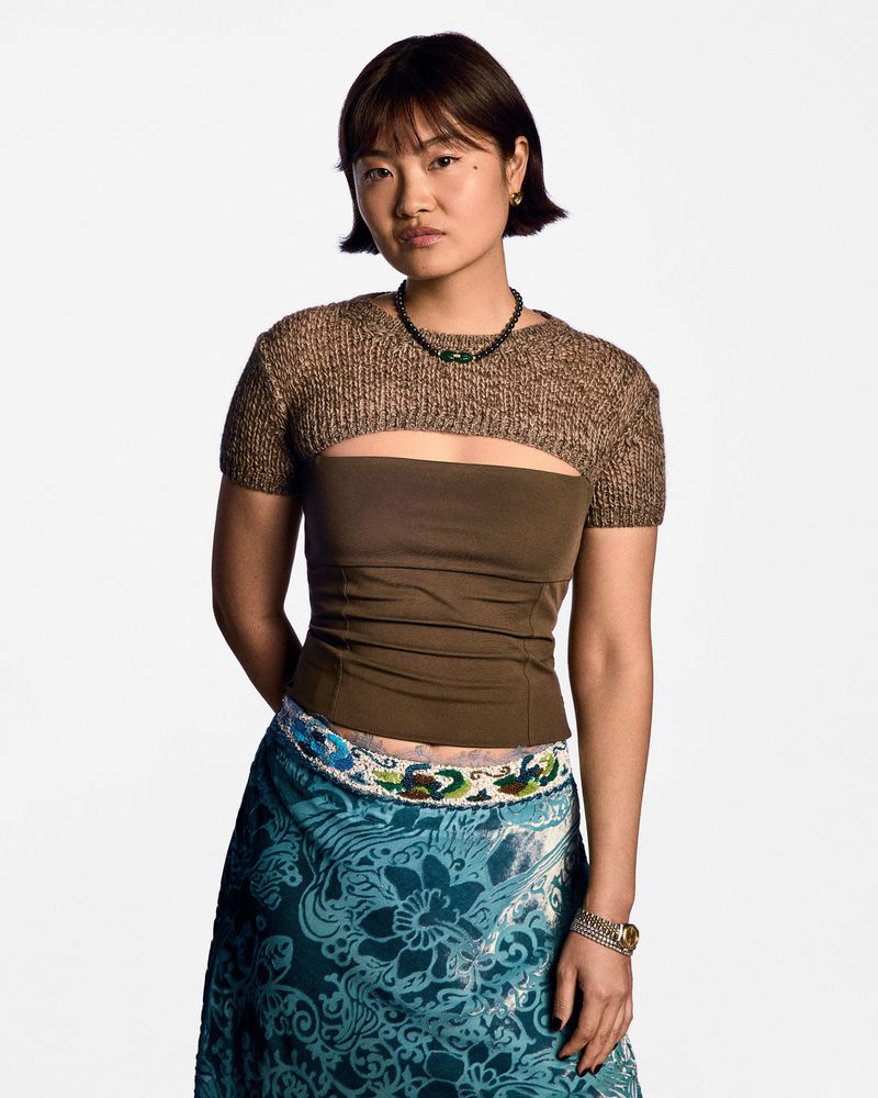Portrait of an Asian woman in brown top and colorful skirt, standing against a white backdrop in a studio setting.