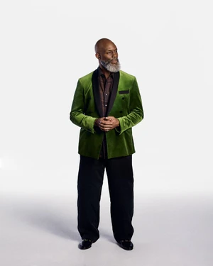 Black man with a grey beard wearing a green velvet blazer and black trousers against a plain white background