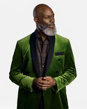 A bald Black man with a grey beard wearing a green velvet blazer over a patterned shirt against a white backdrop.