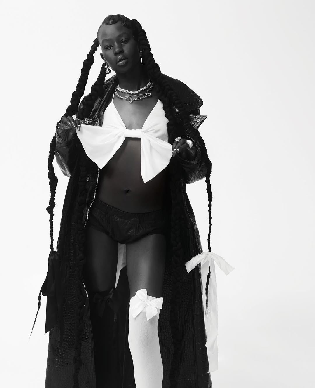 Stylish woman with dark skin and long braids, wearing a bow top and black coat, in a studio shot with bright, even lighting.