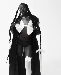 Black woman with long braided hair wearing a dark coat and white bow top against a white background