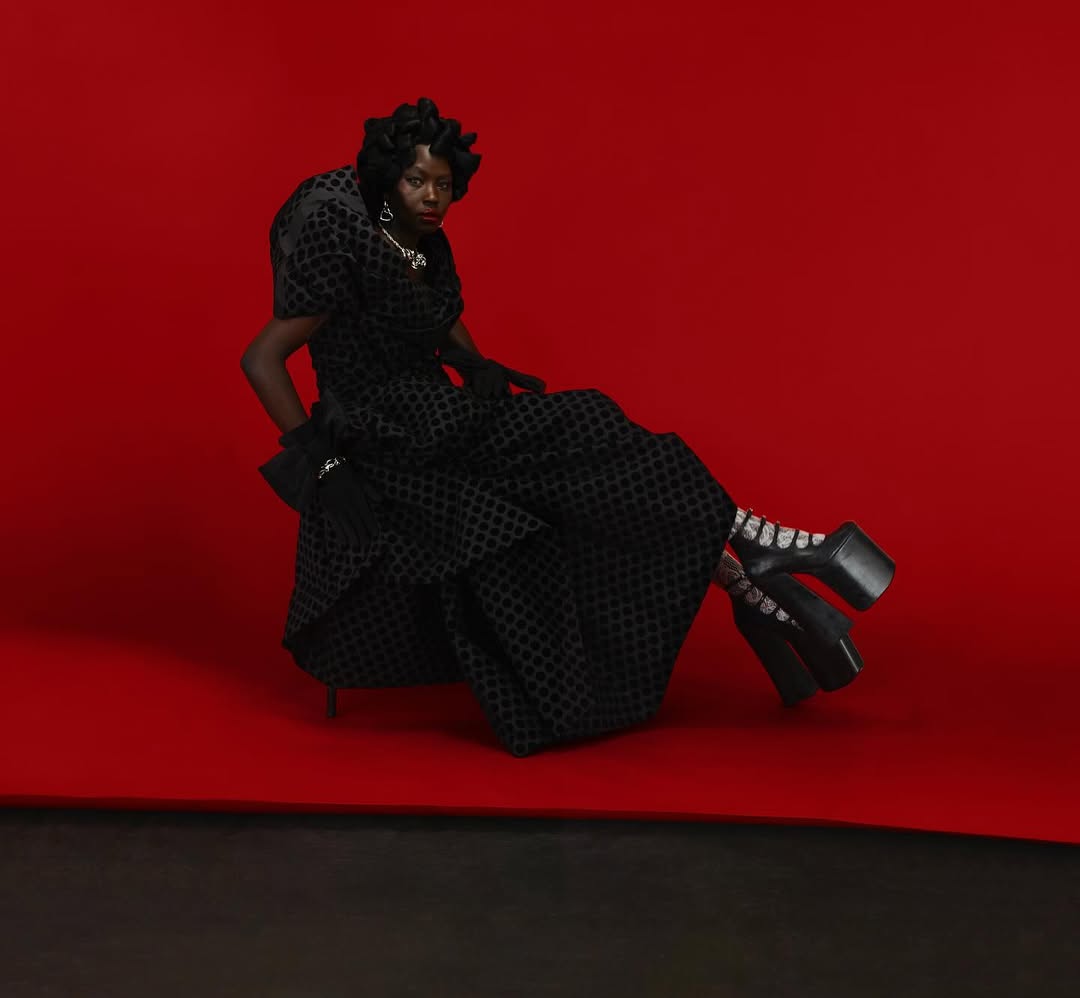 A Black female fashion model poses in a large black gown, jewelry, and platform shoes against a red backdrop in a studio shot.