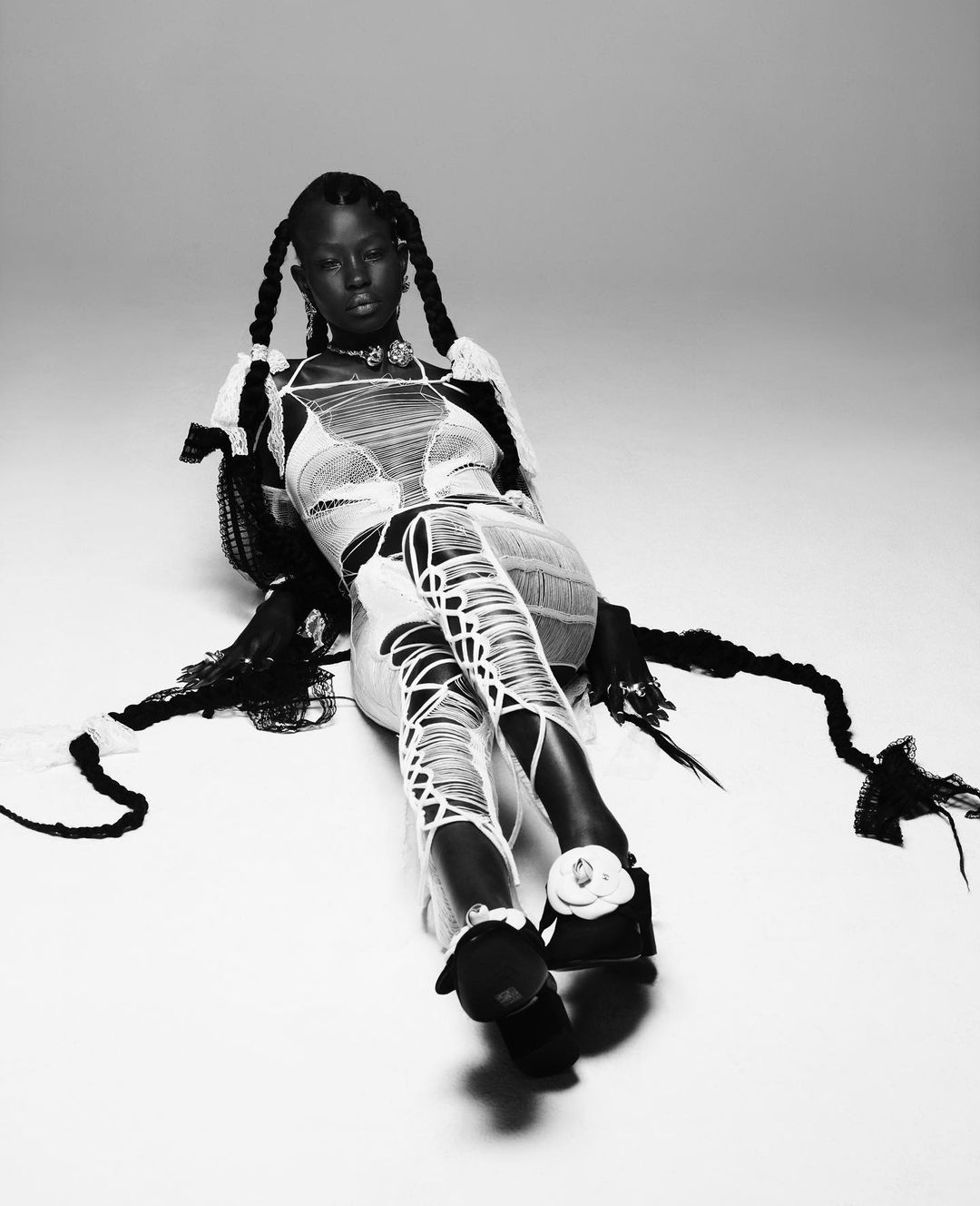 Black-skinned woman in a deconstructed white outfit poses with long braids on a white seamless backdrop in a fashion-style shot.