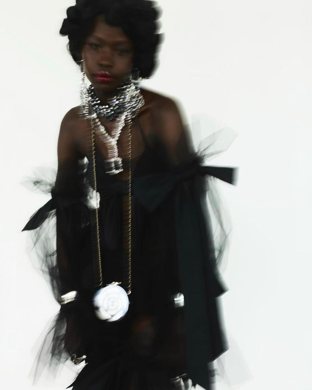 Elegant portrait of a Black woman in a tulle dress with statement jewelry and a purse with a floral design on a white background.