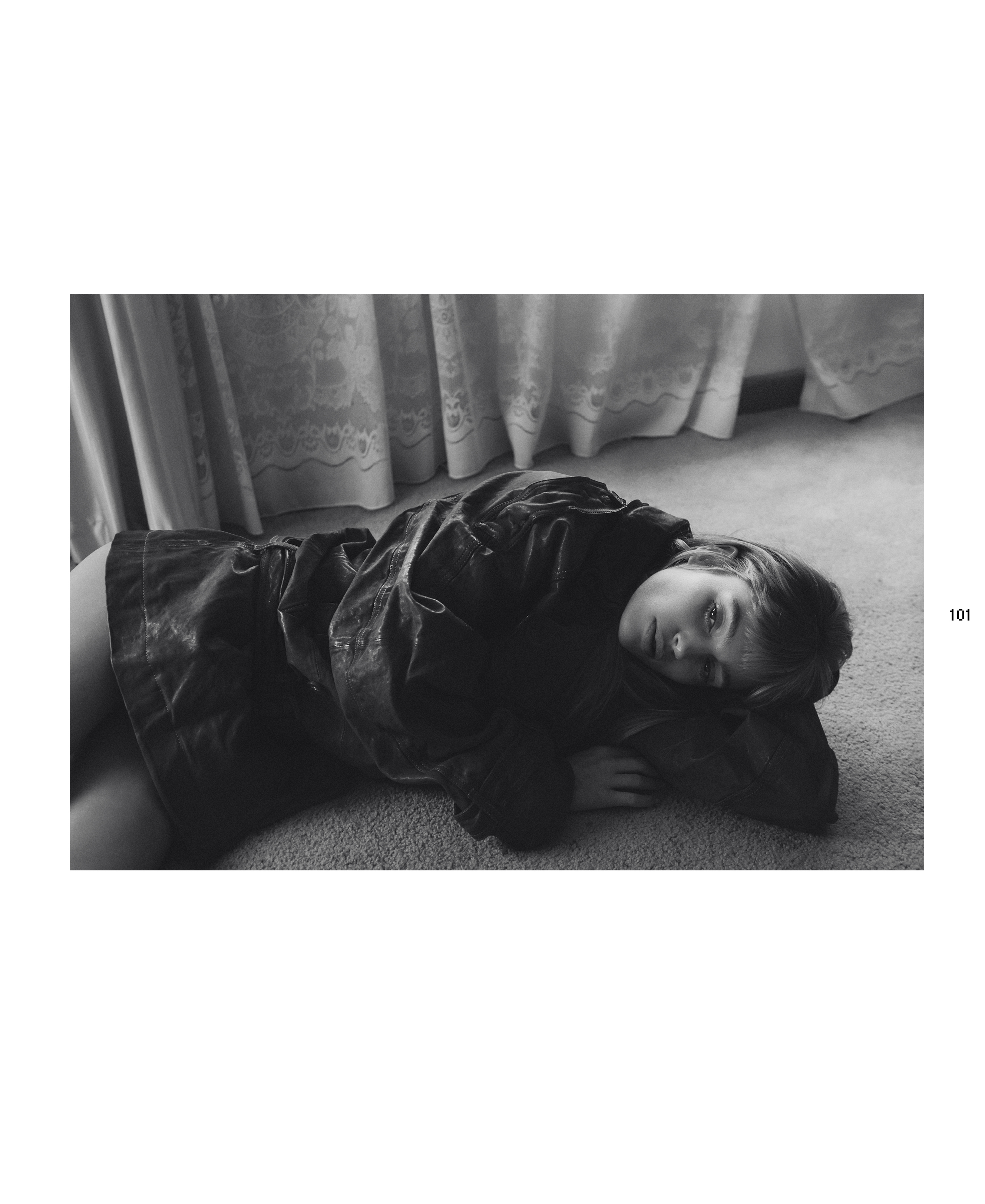 Black and white portrait of a model lying on the floor in a loose jacket with curtains in the background. Editorial fashion photography.