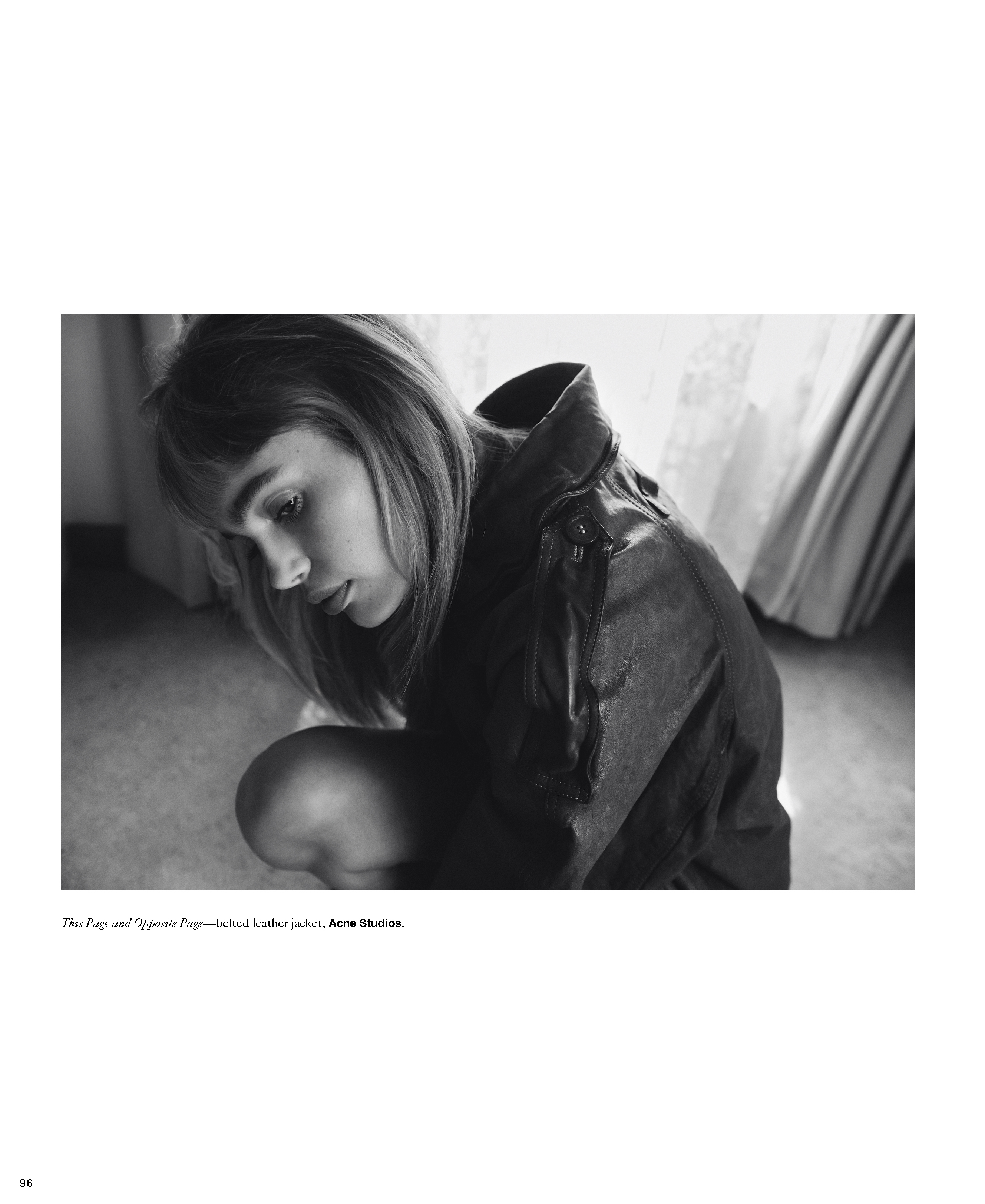 Monochrome close-up of a young woman in a jacket looking down, with a blurred window visible in the background, soft lighting, and editorial style.