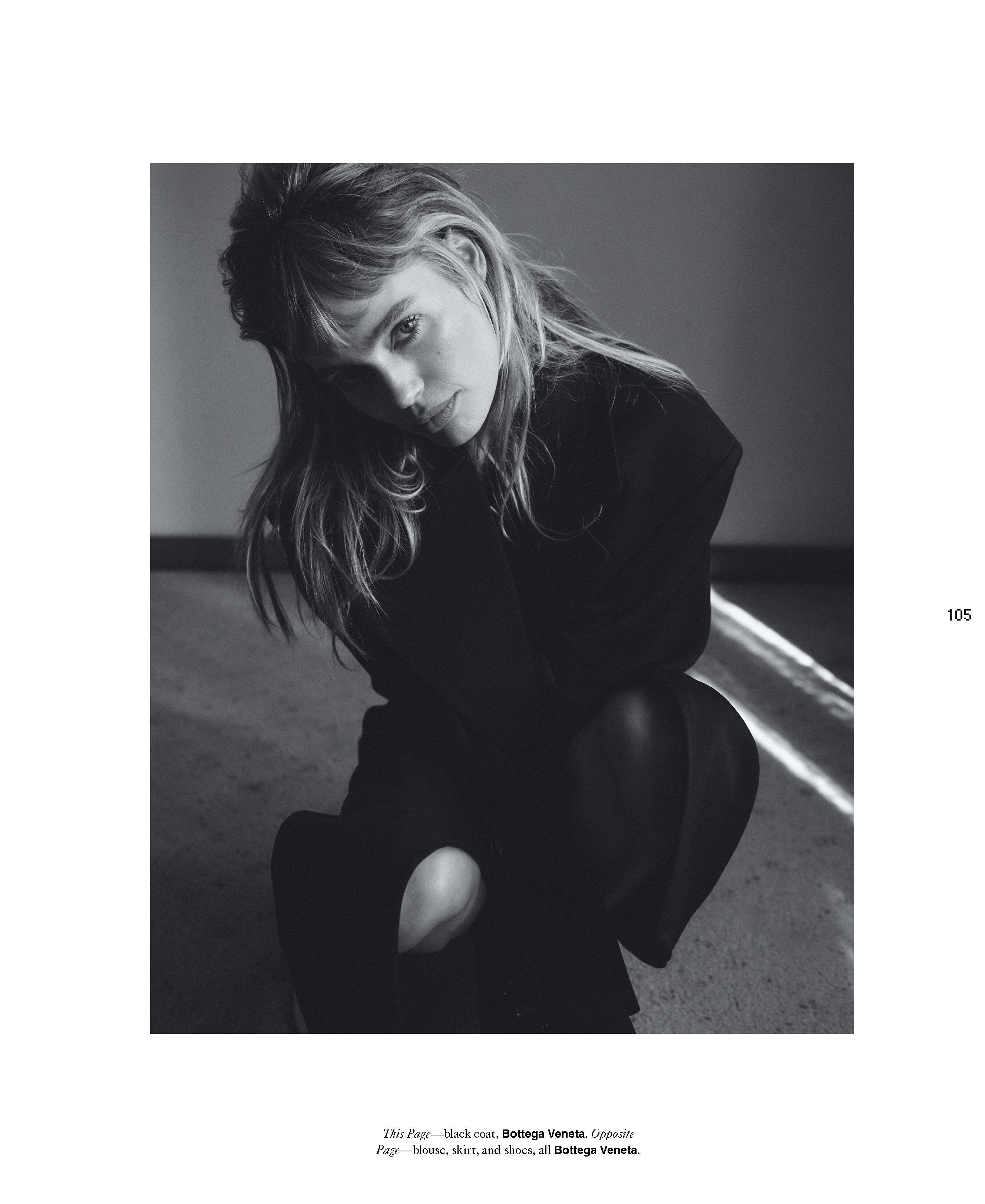 Black and white portrait of a woman with long hair wearing dark clothing, kneeling indoors with natural light streaming across the floor.