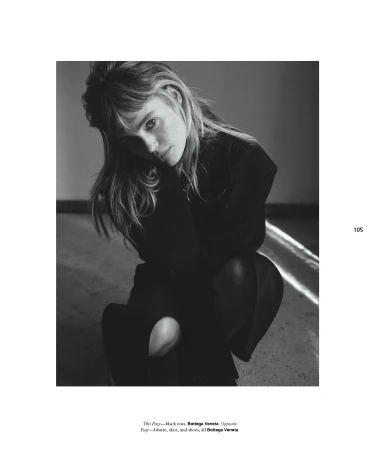 Black and white portrait of a woman with long hair wearing dark clothing, kneeling indoors with natural light streaming across the floor.