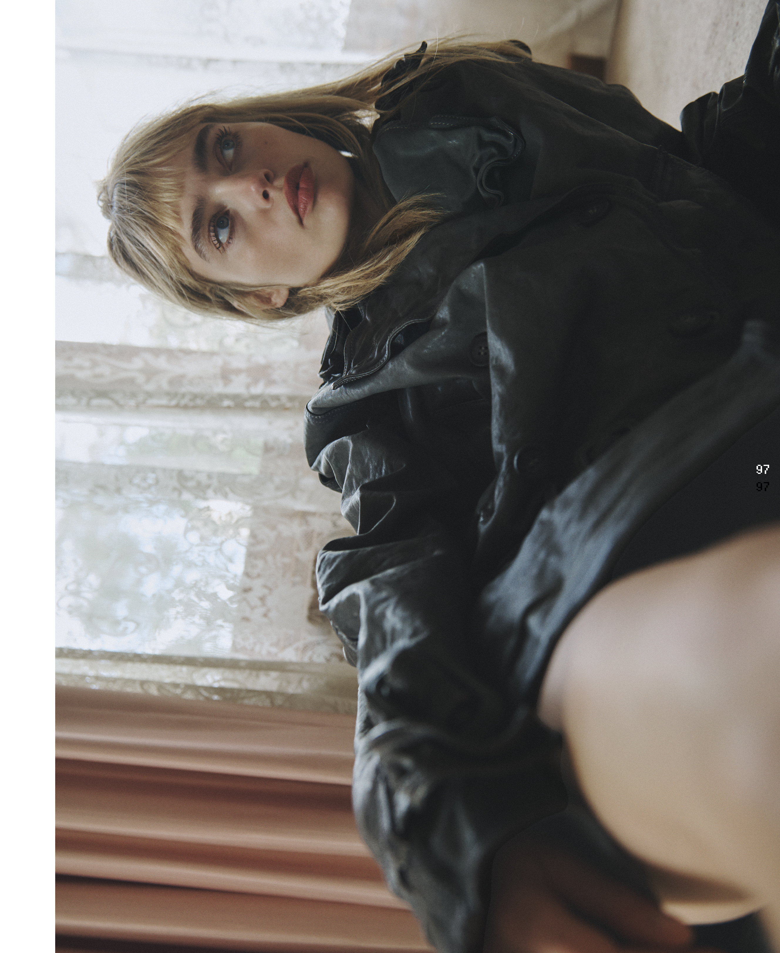 Portrait of a blonde woman in a dark jacket looking up at the camera with a neutral expression; window and curtain in the background.