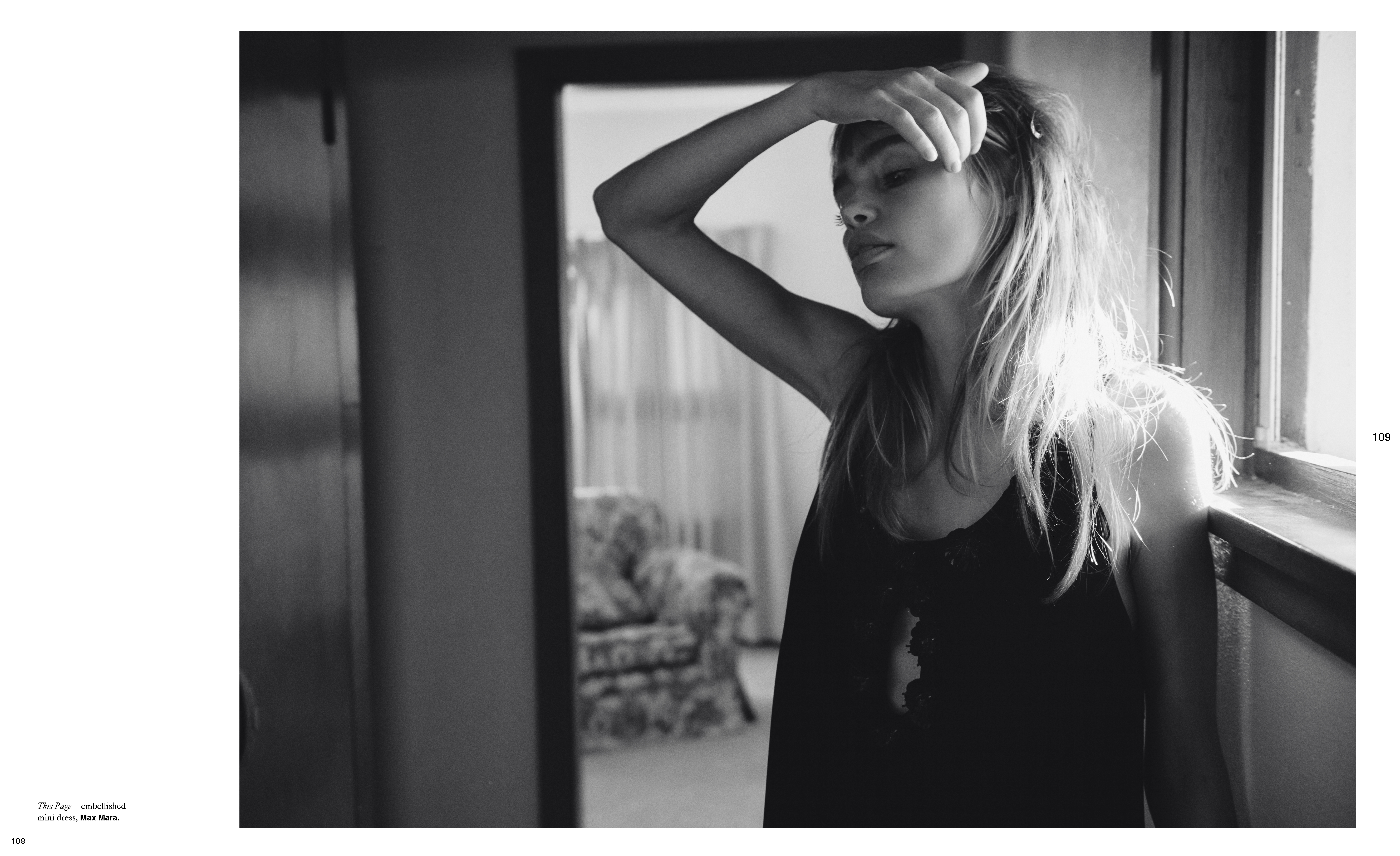 Monochrome portrait of a young woman with her hand on her head, standing by a window in a room, with a doorway in the background.
