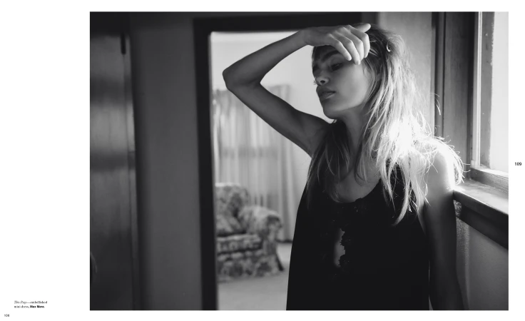 Monochrome portrait of a young woman with her hand on her head, standing by a window in a room, with a doorway in the background.