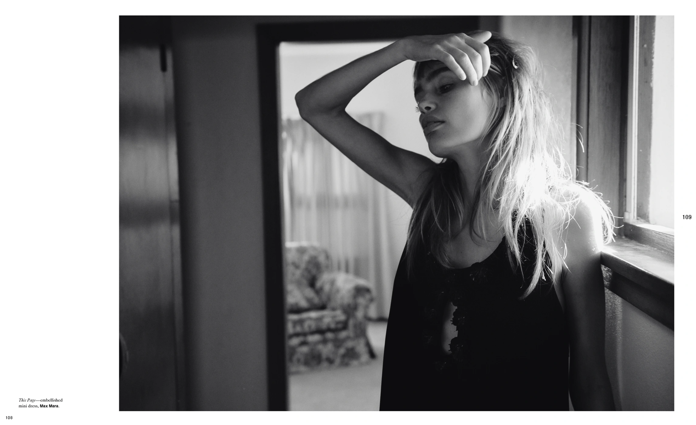Black and white editorial portrait of a woman standing by a window with her hand raised to her forehead