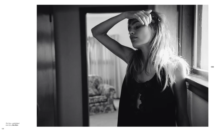Black and white editorial portrait of a woman standing by a window with her hand raised to her forehead