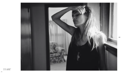 Black and white editorial portrait of a woman standing by a window with her hand raised to her forehead