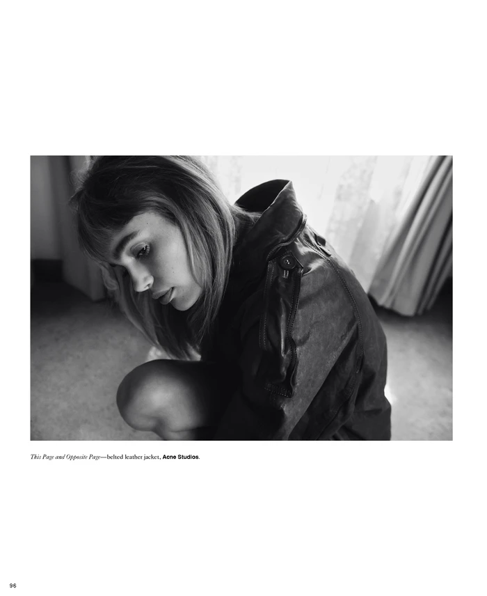 Black and white editorial portrait of a woman crouching in a dark jacket with draped curtains in the background.