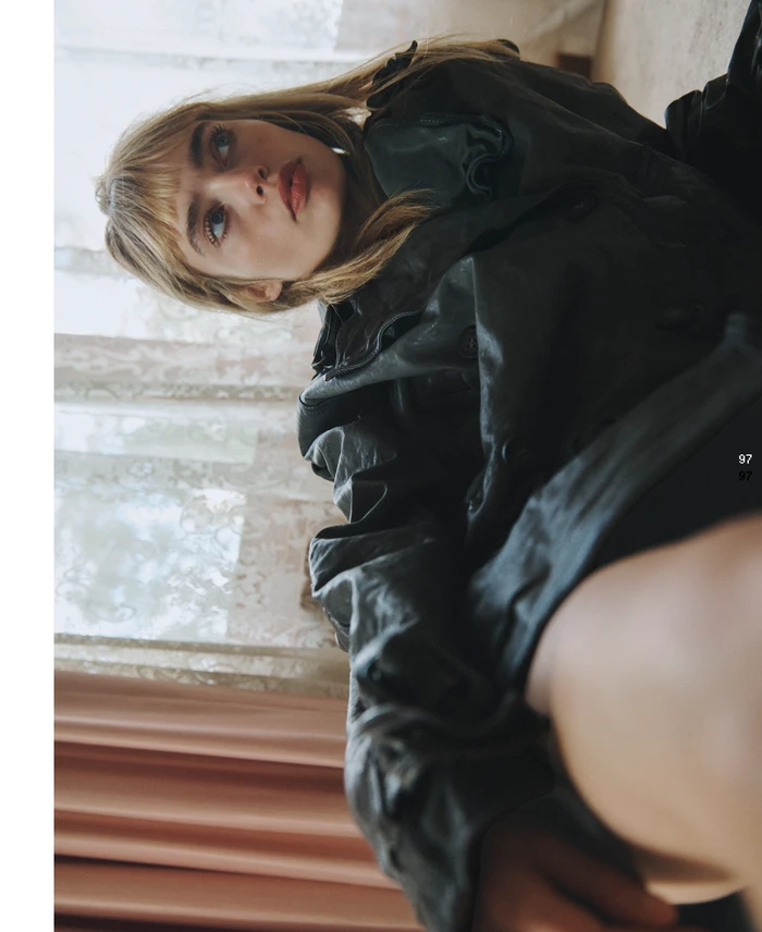 Portrait of a blonde woman wearing a dark coat, captured from a low angle near a window with curtains.