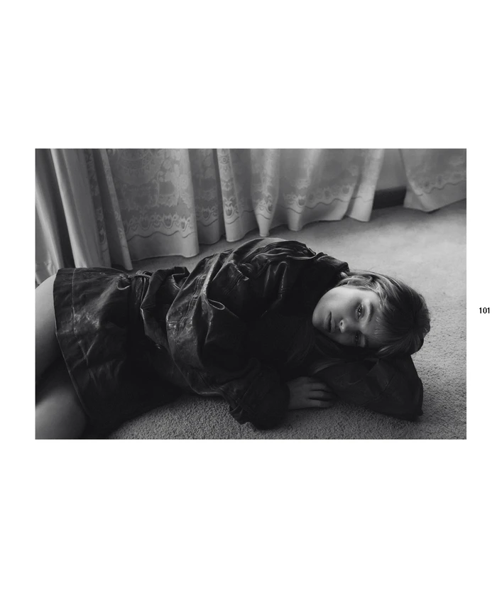 Black and white editorial portrait of a woman lying on carpeted floor wearing a dark jacket with draped curtains behind.