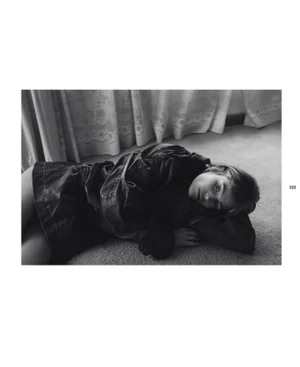 Black and white editorial portrait of a woman lying on carpeted floor wearing a dark jacket with draped curtains behind.