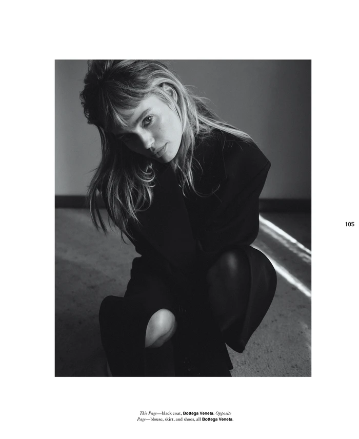 Monochrome fashion portrait of a young woman crouching, wearing a dark top against a plain wall.