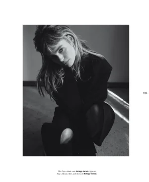 Monochrome fashion portrait of a young woman crouching, wearing a dark top against a plain wall.