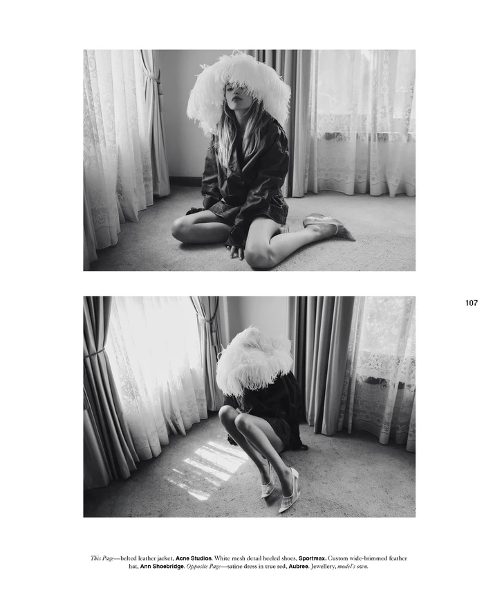 Black and white editorial image of a model wearing a large feathered hat and dark jacket kneeling on a carpeted floor.