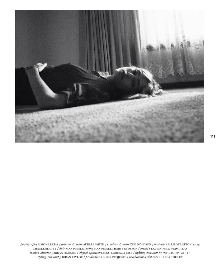 Monochrome editorial portrait of a woman lying on a carpeted floor near a window with sheer curtains.
