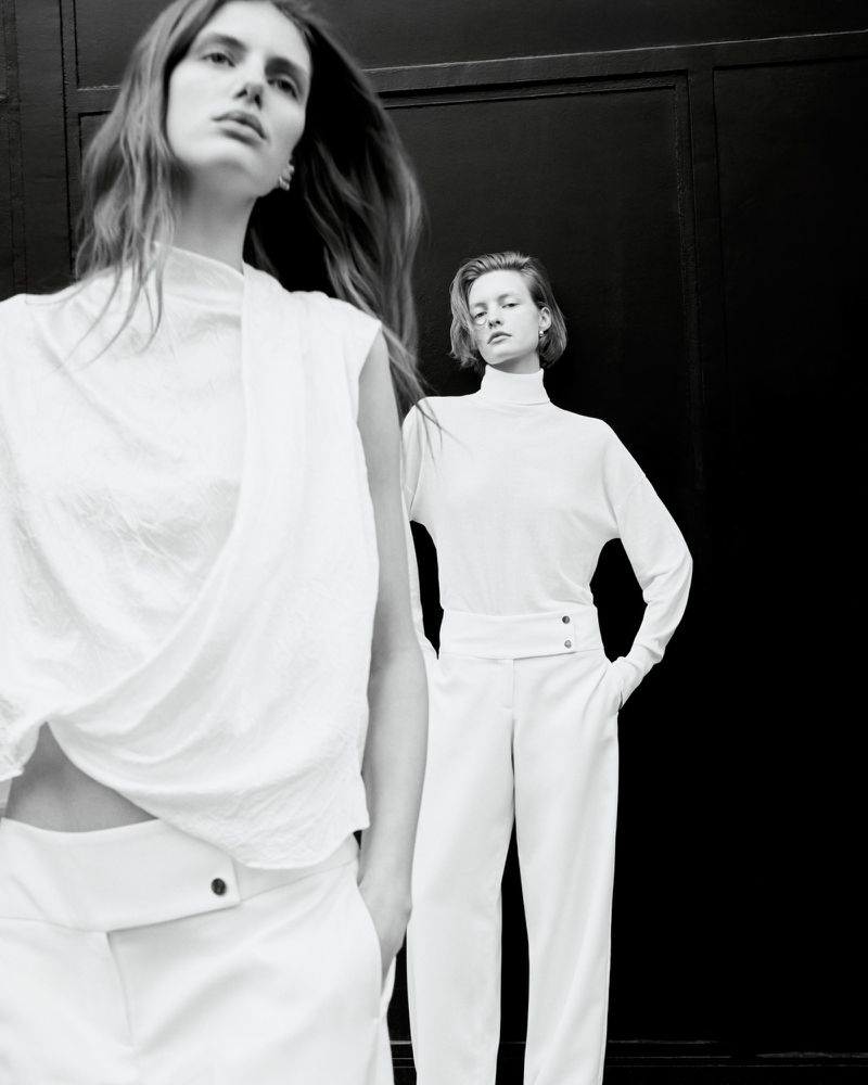 Two adult women pose indoors in white outfits against a dark background, showcasing minimalist fashion and studio photography in monochrome.