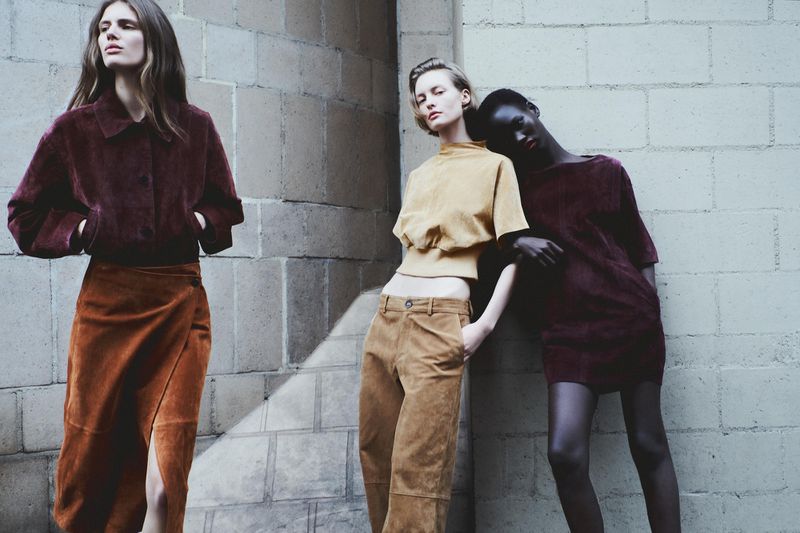 Three fashion models in stylish coordinated outfits pose against a textured wall in a medium outdoor shot, showcasing autumn color palettes.