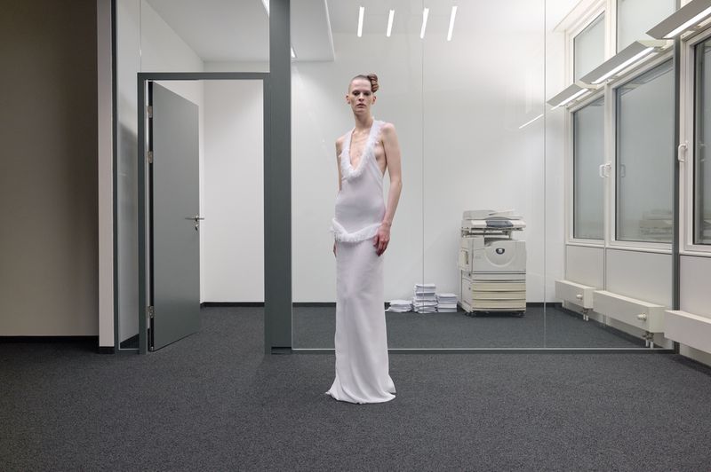 A slender fashion model in a long white dress poses in an office setting with gray carpet, white walls, and a copy machine visible.