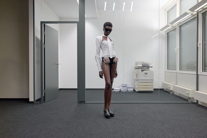 Elegant African American woman poses in an office wearing sunglasses, a white top and black underwear with roses, and platform shoes.