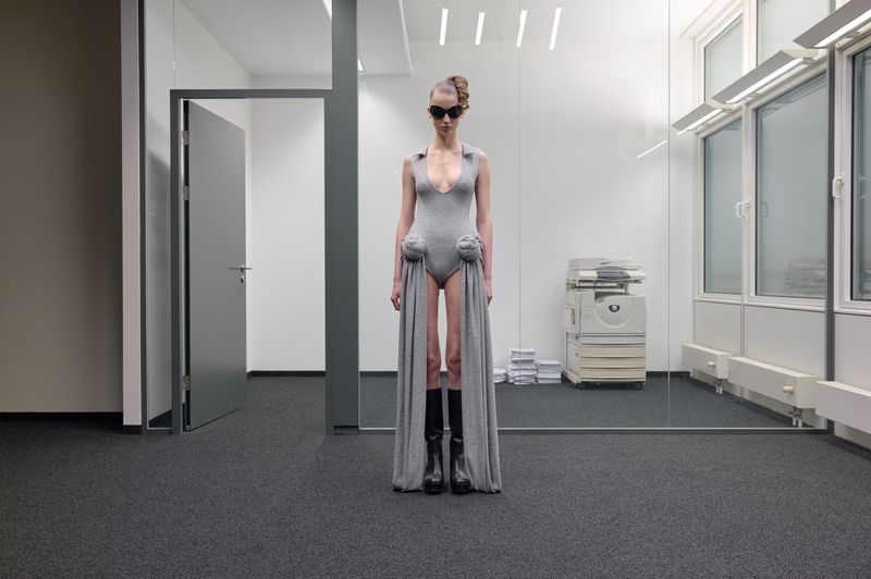 Woman in gray bodysuit and black boots stands in an office space with copy machine and rows of windows with lighting fixtures.