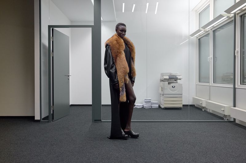 A fashionable woman stands in an office interior wearing a fur stole and a black dress with windows and a printer in the background.