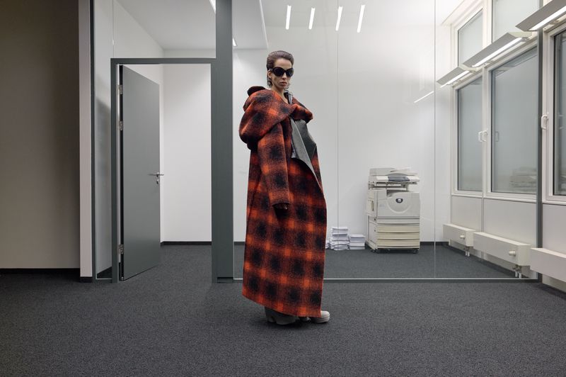 Fashion model in a red and black plaid hooded coat stands in a grey office with white walls, large windows, and a copy machine.